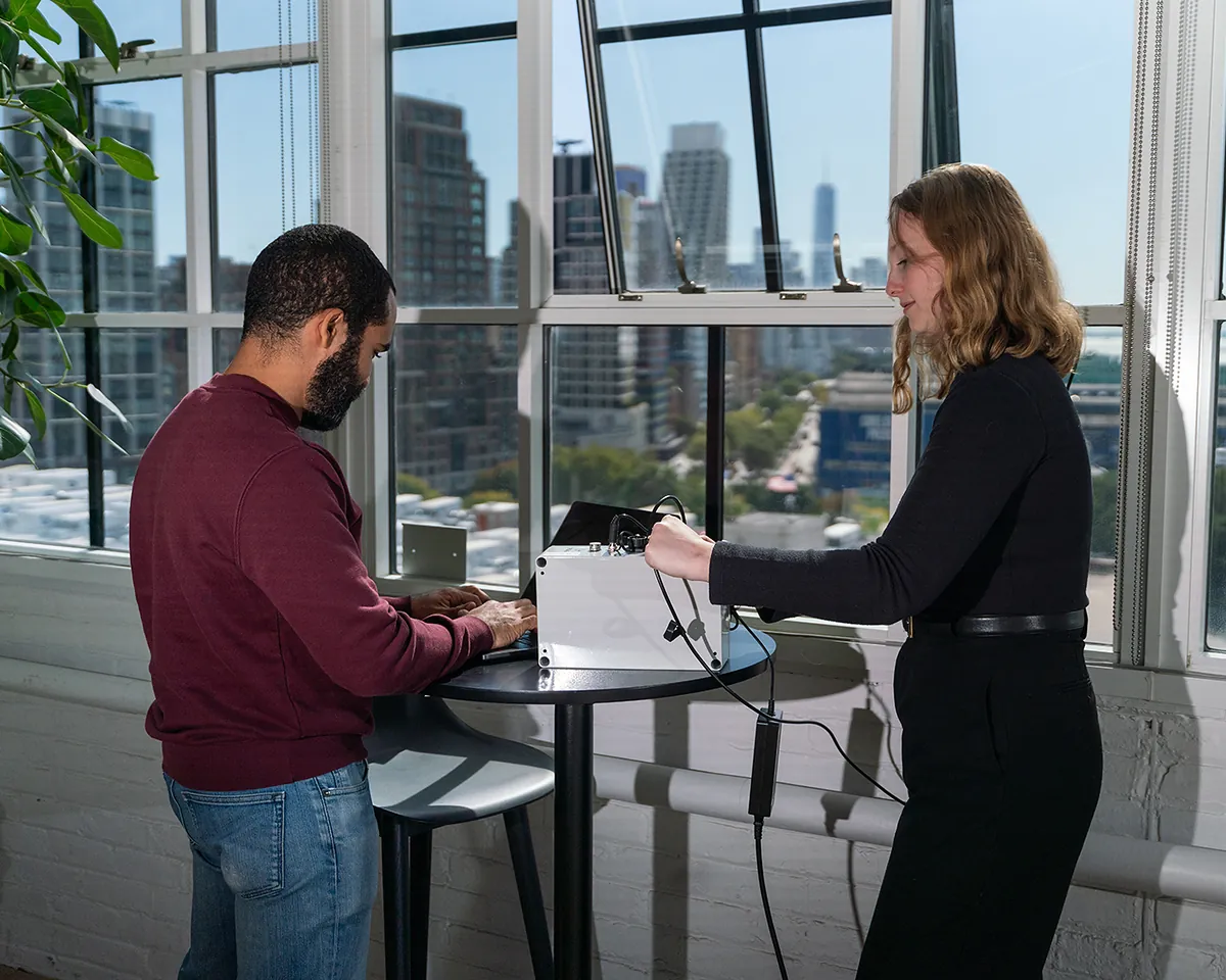 Two team members testing a sensor at a table by the windows, one typing on a laptop while the other connects a cable, with Lower Manhattan behind them